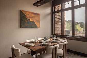 A dining area at Alpenglow Condo 26D featuring a set wooden table for six, an abstract painting, and large windows with mountain views.