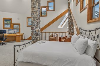 A spacious bedroom featuring a large bed with white linens, an office desk with two monitors, and a cozy reading nook with two armchairs, all set against a backdrop of stone and wood architectural details.