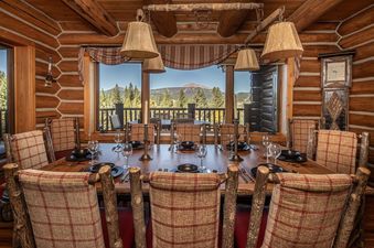 A rustic dining room with a large wooden table and chairs, offering a scenic view of mountains and forests through expansive windows.