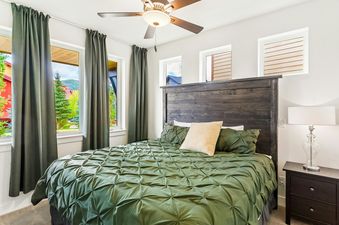 A bedroom with a green comforter, a wooden headboard, and two windows with curtains.