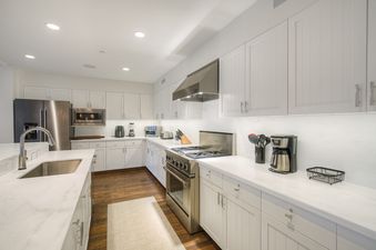 A modern kitchen with white cabinets, marble countertops, and stainless steel appliances, including a refrigerator, microwave, stove, and sink. Dark wood floors are visible beneath.