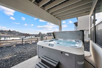 A hot tub with its cover partially open, showing bubbling water, sits on a patio with a scenic view of a lake, playground, and residential area under a blue sky.
