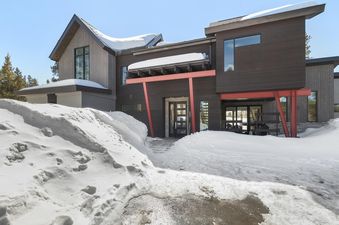 The Modern Alpine Escape is a contemporary dark wood and red-accented home with large windows, surrounded by deep snow under a clear winter sky.