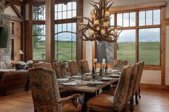 Rustic dining table and antler chandelier in the Hord Mountain Retreat, with scenic window views of green fields and distant mountains.