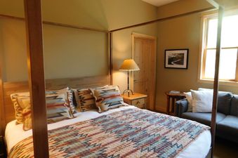 A cozy bedroom features a king four-poster bed with a colorful quilt, a nightstand with a lamp, a gray couch, and a large window.