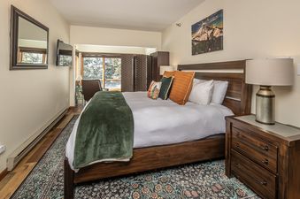 The bedroom at Snowflake at Skycrest features a queen-sized bed with a wooden headboard, a white duvet, and a green throw blanket, complemented by a rustic nightstand, a mountain artwork, and a large window with a snowy outdoor view.