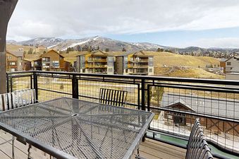 A balcony with a mesh table offers a stunning view of a mountainous landscape with buildings and a clear sky.