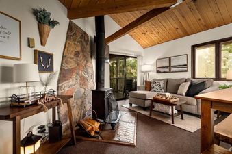 A cozy living room at Cedar Creek Cabin featuring a stone fireplace, wood beam ceilings, a comfortable sectional sofa, and large windows with outdoor views.