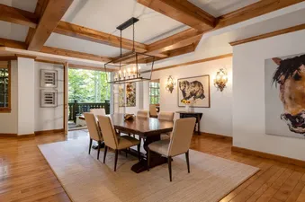A luxurious dining room with a long wooden table, cream chairs, exposed wooden beams, and a view of green trees through large windows.