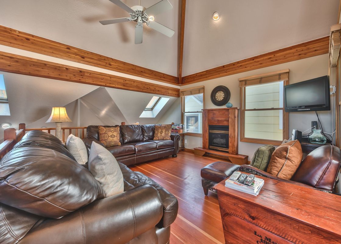 A cozy living room with leather sofas, a wooden coffee table, a fireplace, and a television, all under a vaulted ceiling with wooden beams.