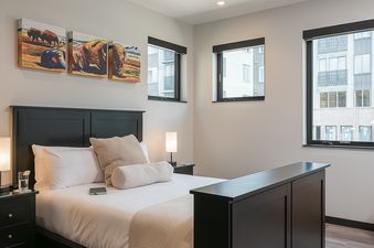 The bedroom in Triple 3 Lodge offers a serene retreat with a comfortable bed, nightstands, and captivating wall art, illuminated by natural light from multiple windows.