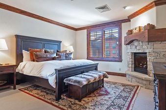 A cozy bedroom featuring a large bed, fireplace, and window with wooden shutters.
