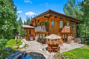 An expansive wooden mountain home, the Lupine Estate, is nestled amidst vibrant green trees, featuring a large patio with outdoor dining, a grill, and a built-in hot tub under a bright blue sky.