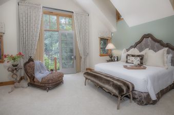 The luxurious master bedroom at Pine Meadow Estate boasts a plush king-sized bed, a cozy armchair, and a gorgeous view of the surrounding woods from the large windows.