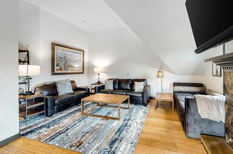 A cozy living room featuring dark leather sofas, a patterned area rug, a wooden coffee table, and various lamps, with a framed ski resort map on the wall.