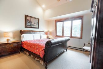 A spacious bedroom features a large, ornate wooden sleigh bed with a red and white patterned comforter, flanked by a wooden nightstand and a floor lamp, with a large window offering a view of lush trees outside.