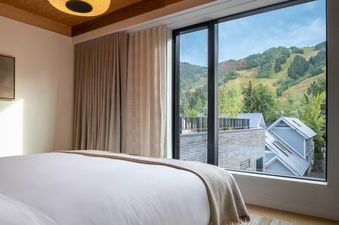 A modern bedroom at Mollie Aspen Mountain King features a king-size bed facing a large window with views of green mountains and residential rooftops.