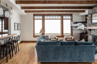 The living room features a large, comfortable sofa, a stone fireplace, and windows with views of the snowy mountain peaks.