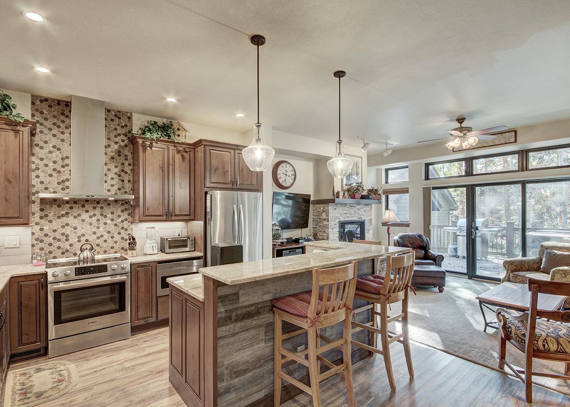 A modern kitchen with a stainless steel stove and a breakfast bar that overlooks a living room with a fireplace and large windows.