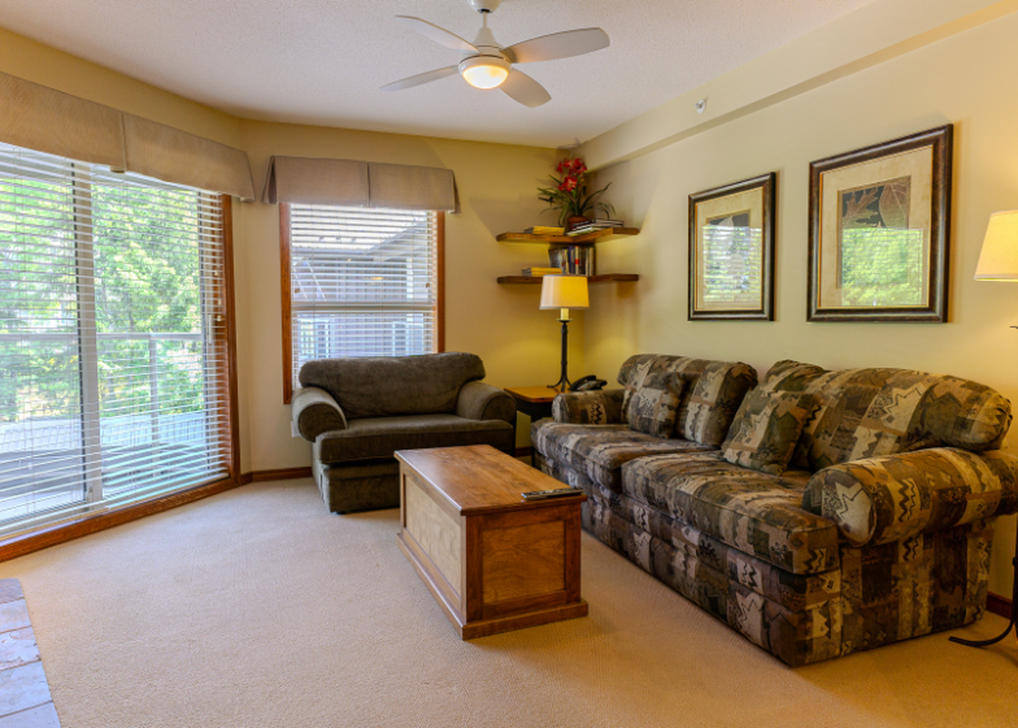 A cozy living room in Aspens on Blackcomb Unit 438 features a patterned sofa, a matching armchair, and a wooden coffee table, all bathed in natural light from large windows offering a lush green exterior view.