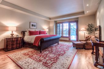 A luxurious bedroom in Cowboy Heaven Luxury Suite Unit 2B features a large bed with red accents, a cozy armchair, and a window with a view of a snowy landscape.