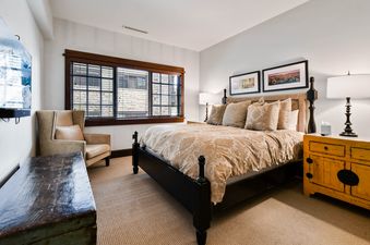 A cozy bedroom in Arrowleaf Lodge 302 featuring a king-sized bed with patterned bedding, two lamps, wall art, a window with blinds, an armchair, and a rustic wooden chest.