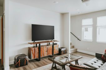 A TV sits on a sleek wooden cabinet in The Circle Unit B's bright living room, complemented by a glass coffee table and a dark leather sofa.
