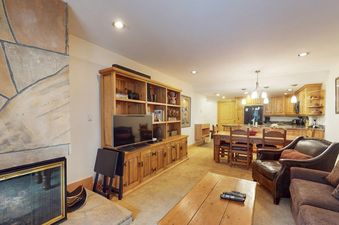 The open-concept living and dining area in Fallridge 402, featuring a stone fireplace, a large wooden entertainment center with a TV, a dining table, and a kitchen in the background.