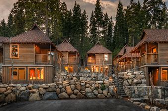 A row of wooden cottages with glowing windows and outdoor string lights are nestled among tall pine trees, featuring stone retaining walls and staircases leading to private entrances.