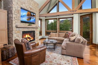 The living room in Pines Townhome 10 features a stone fireplace with a TV above, a sectional sofa, a leather armchair, a wooden coffee table, and large windows revealing an outdoor view.