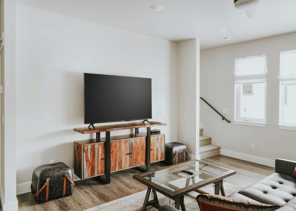 A TV sits on a sleek wooden cabinet in The Circle Unit B's bright living room, complemented by a glass coffee table and a dark leather sofa.