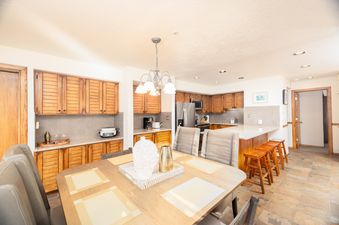 The Snow Park Chalet features an open-concept dining area and kitchen, furnished with a wooden dining table, upholstered chairs, and a breakfast bar with wooden stools, all set against a backdrop of modern appliances and warm wooden cabinetry.