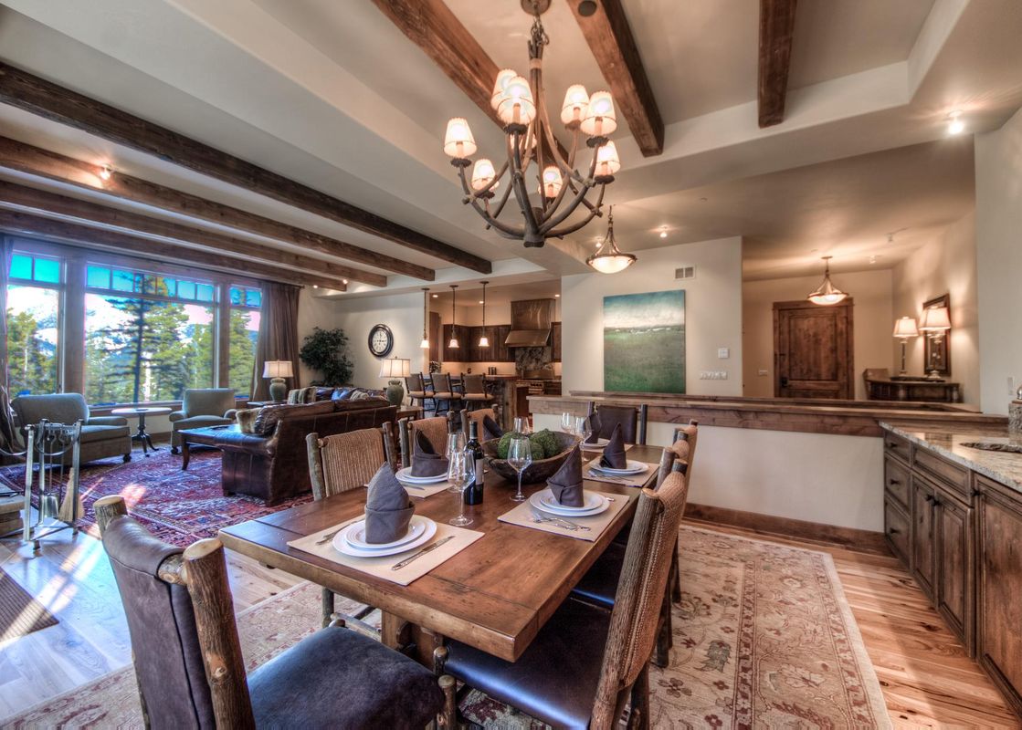 A luxurious dining and living area with rustic wooden furniture, exposed beams, and a large window overlooking a forest.