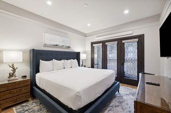 A sophisticated bedroom in The Charter D3105 featuring a king-size bed with a navy blue headboard, two nightstands with unique lamps, and a door with decorative glass leading to an outdoor area.