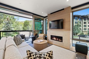 A modern living room at Assay Hill Lodge Unit 264 with a cozy fireplace, large sectional sofa, and expansive windows offering stunning mountain and architectural views.