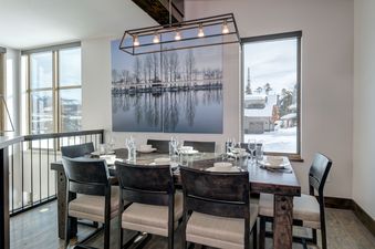A spacious dining area at Homestead Chalet 8 Claim Jumper with a large dark wood table set for a meal, illuminated by a modern chandelier, and offering scenic window views of a snowy landscape.