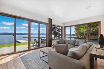 A modern living room with a large grey sectional sofa and a wooden coffee table, featuring floor-to-ceiling windows that offer an expansive view of a lake, mountains, and a bright blue sky.
