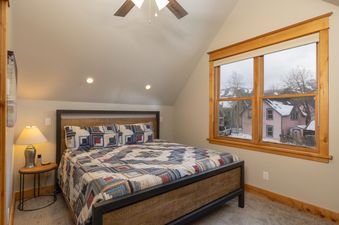 A cozy bedroom with a patterned bedspread, a bedside table with a lamp, and a large window offering a view of snowy houses and trees.