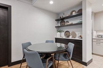 A dining area with a round table and four gray chairs is depicted, complemented by floating shelves with decor and a partial view of a kitchen.