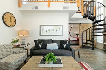 A modern rustic living room with a gray sofa, antler-print pillows, a wooden coffee table, and a black spiral staircase. A framed artwork of deer hangs above the sofa.