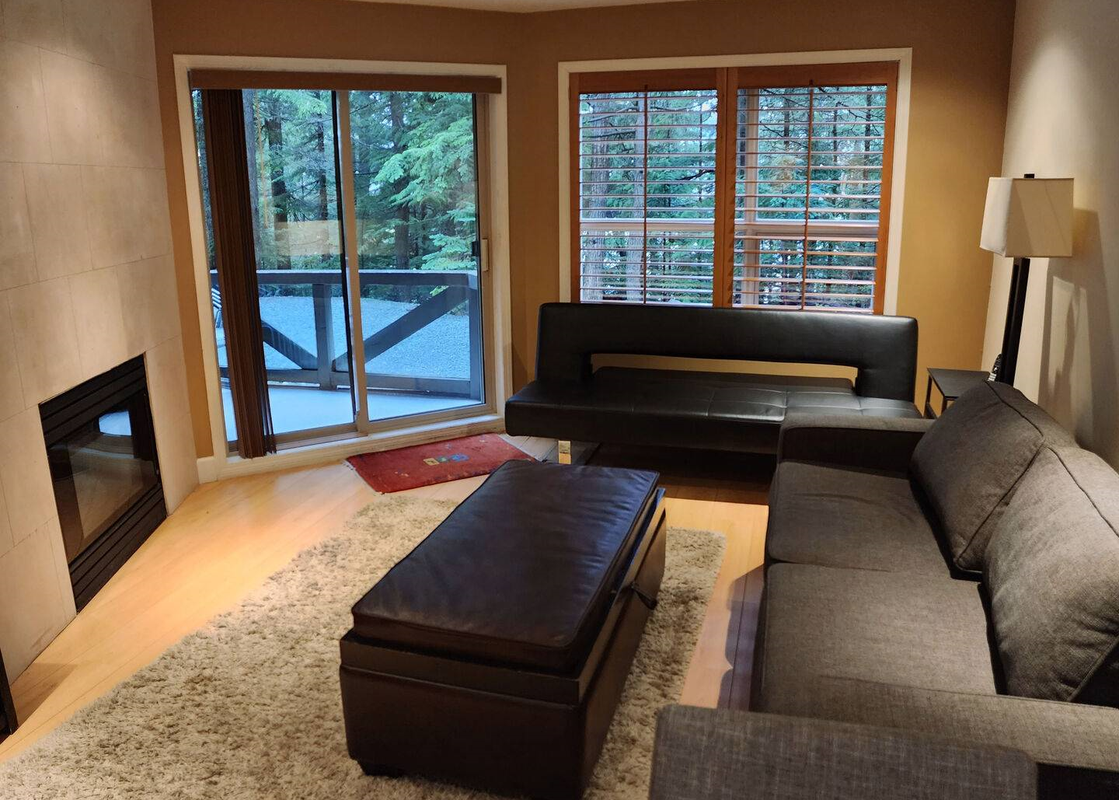 The Aspens on Blackcomb Unit 113 features a cozy living room with a large gray sectional sofa, a dark brown leather ottoman, and a fireplace, offering a comfortable and inviting atmosphere.