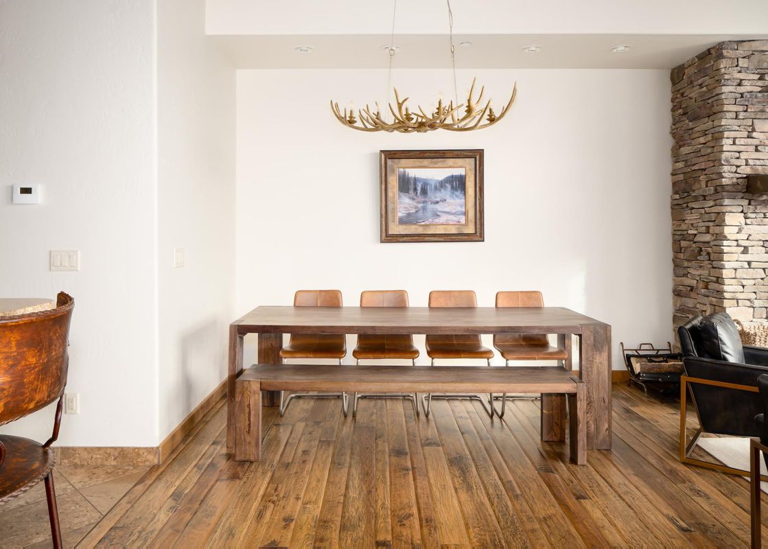A dining area with a dark wood table, leather chairs, and a bench, illuminated by an antler chandelier, with a framed landscape painting and a stone fireplace nearby.