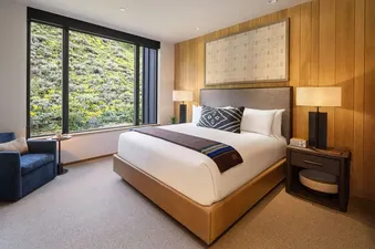 A modern bedroom in The Sylvan Lodge One Bedroom Junior Penthouse features a king-size bed, a wooden accent wall, and floor-to-ceiling windows with views of a lush, green mountain landscape.