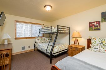 The bedroom at Krystal Villa 5 features a bunk bed with colorful bird-patterned bedding, a wooden nightstand with a lamp, and a desk with a lamp, all set on a carpeted floor with a large window in the background.