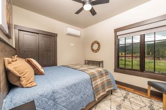 A cozy bedroom at Sky High Colorado features a bed with a blue quilt and plaid throw, a rustic headboard, a dark wood closet, an air conditioning unit, a round mirror above a console table, and a large window with a scenic view of green hills and mountain