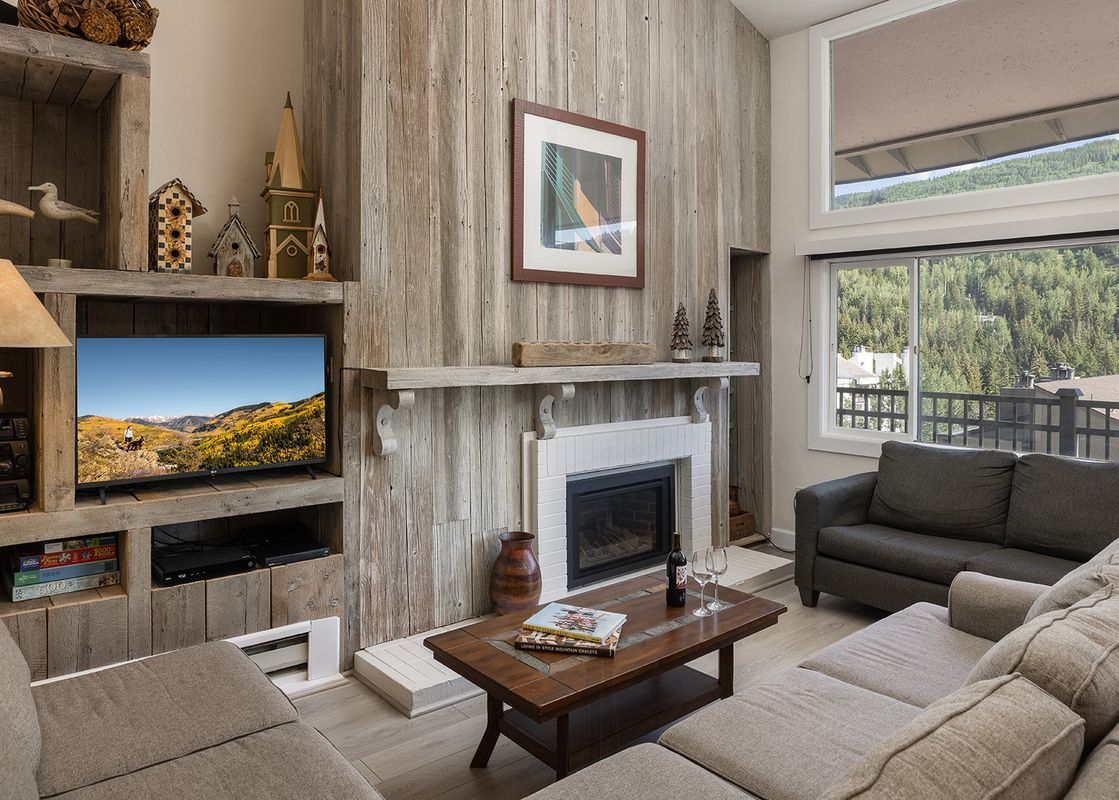 A cozy living room at Vantage Point 501 with two couches, a television, a fireplace, and large windows overlooking a scenic mountain and forest view.