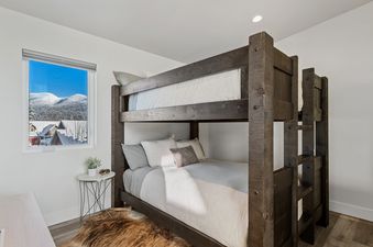 A bunk room at Quarry A28 features a rustic wooden bunk bed with neutral bedding, a small side table, and a window with a view of snowy mountains and rooftops.