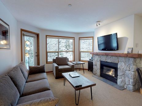 A cozy living room in Aspens on Blackcomb Unit 443 with a stone fireplace, comfortable sofa, armchair, and views of a snow-covered landscape.