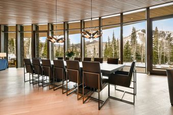 The dining room at Iron Mountain Chalet features a long table with modern chairs, surrounded by floor-to-ceiling windows offering a panoramic view of a snow-covered mountain landscape with evergreen and aspen trees.