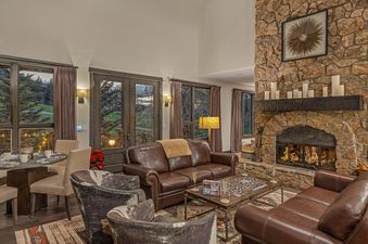 The Aspen Glade Retreat features a spacious living area with a stone fireplace, comfortable leather sofas, and a dining table, all illuminated by large windows overlooking a lush landscape.